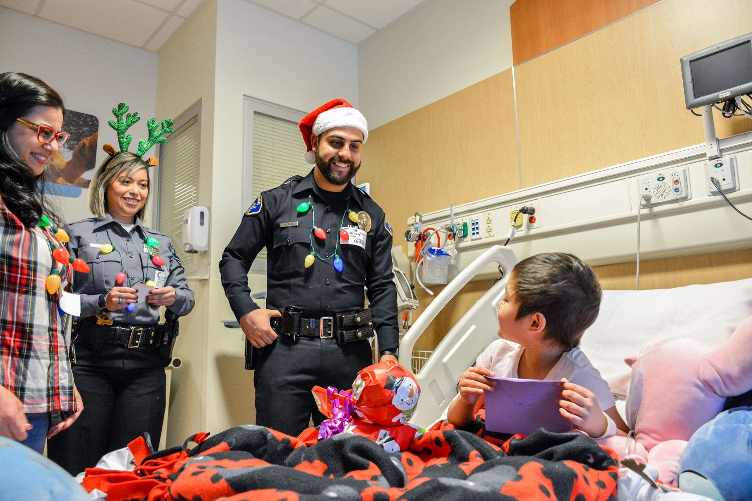 Police officers and a volunteer smile at a child opening a present in a hospital room